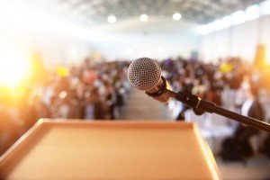 Standing at a podium with a microphone facing the camera, looking out at the audience