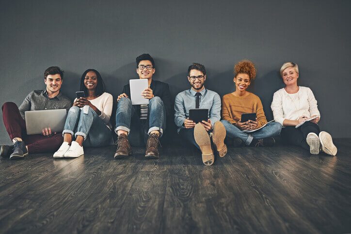 A group of millenials sitting on the floor, against a wall with devices in their hands