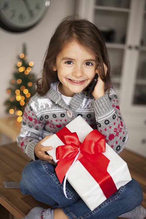 Young girl talking on phone holding a present in front of a christmas tree